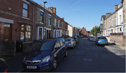 A narrow Victorian terraced street with cars parked partly on the pavement on both sides
