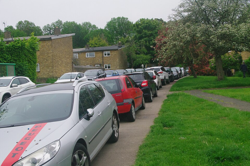 Cars parked with two wheels on a grass verge and pavement outside 1950s housing, with a footpath alongside